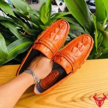 Hình đôi giày tây GCA2 da cá sấu gai đuôi màu vàng cam trên chân, nền lá xanh, thể hiện thời trang cao cấp đẳng cấp quý ông.