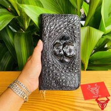 Hình ảnh bàn tay cầm ví da cá sấu đen với họa tiết gù lồi, nền lá xanh và thẻ logo.