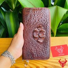 Hình ảnh bàn tay cầm ví da cá sấu nâu đất với họa tiết gù lồi, nền lá xanh và thẻ logo.